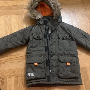 Grön vinterjacka med fuskpäls från BLWR - Mörkgrön pufferjacka från BLWR med huva och fuskpäls, orange detaljer och flera stora fickor framtill. Jackan har dragkedja och tryckknappar, samt ett orangefärgat innerfoder. Perfekt för kalla dagar!