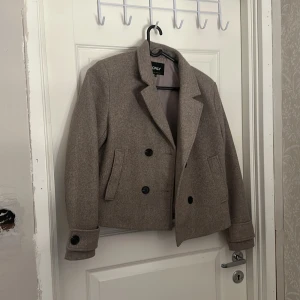 Beige skepparkavaj från ONLY, stl M - Snygg beige skepparkavaj från ONLY i klassisk modell med dubbelknäppning och krage. Jackan har raka fickor framtill, långa ärmar med knappdetalj vid ärmslut och är tillverkad i ett ull-liknande material. Perfekt för dig som gillar stilrena och tidlösa plagg. OBS!! Endast testad.