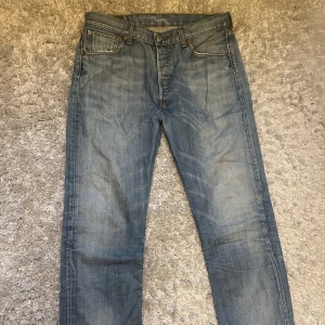 Levi's 501 ljusblå jeans straight fit - Klassiska Levi's 501 jeans i ljusblå tvätt med raka ben och normal passform. Jeansen har fem fickor, knappgylf och tydliga kontrastsömmar. Tillverkade i slitstarkt bomullsjeansmaterial och har den ikoniska läderpatchen bak i midjan.