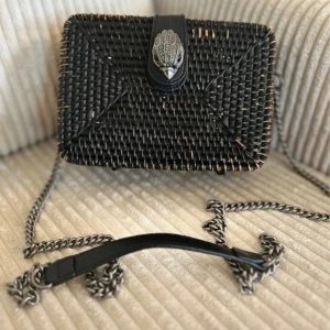 Svart axelväska från Kurt Geiger - Snygg svart axelväska från Kurt Geiger i flätad design med silverfärgad kedjerem och coolt örndekor på locket. Väskan är ca 18,5 cm bred och har svart innerfoder med logga. Perfekt storlek för det viktigaste och ger en trendig touch till din outfit.