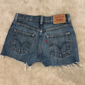  Låga Levi's jeansshorts med fransig kant - Snygga blå jeansshorts från Levi's med klassisk femficksdesign och fransig, rå kant nedtill. Modellen har normal passform och low waist. Köpte de second hand men är i väldigt bra skick. Måste tyvärr sälja då de blivit försmå. Hör av dig för fler bilder! 