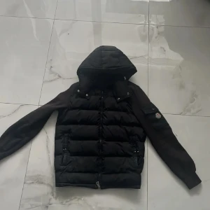 Svart pufferjacka från Moncler - Svart pufferjacka från Moncler med huva och stickade ärmar. Jackan har dragkedja framtill, två fickor med knappar och en extra ficka på ärmen med Moncler-logga. Perfekt för kyliga dagar och riktigt snygg streetstil.