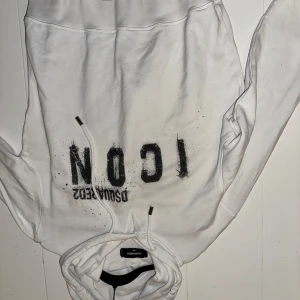 Vit ICON hoodie från Dsquared2 - Säljer en vit hoodie från Dsquared2 med svart 'ICON' print och coola färgstänk på framsidan. Tröjan har huva med snörning och långa ärmar. Perfekt för dig som gillar streetwear och vill sticka ut med en snygg designertröja.