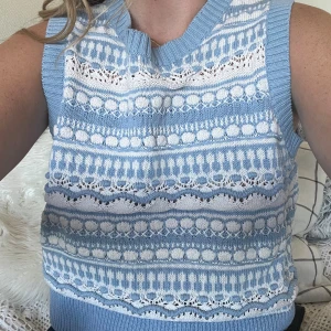 Blåvit stickad väst  - Superfin stickad väst i ljusblått och vitt med ett randigt och mönstrat design. Västen har ribbade kanter vid hals och ärmhål, samt coola detaljer med vågiga och prickiga inslag. Perfekt att styla över en t-shirt eller skjorta för en trendig look.