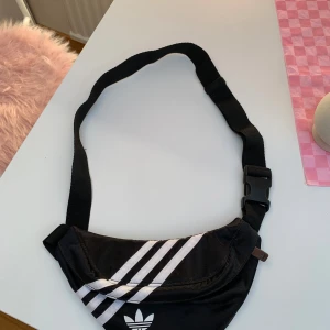 Svart axelväska från Adidas - Säljer en svart axelväska från Adidas med klassiska vita ränder och logga framtill. Väskan har justerbart band och smidig plastspänne. Perfekt för att bära det viktigaste och passar till streetwear-stilen.