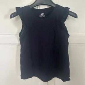 Svart ärmlös topp med volang H&M - Svart ärmlös topp från H&M i mjuk bomull. Toppen har rund halsringning och volangdetaljer vid axlarna som ger en söt och trendig look. Perfekt att matcha med jeans eller kjol för en enkel och stilren outfit.