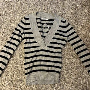 Randig v-ringad tröja från Gina Tricot - Säljer en grå och svart randig tröja från Gina Tricot med djup v-ringning och ribbade muddar. Tröjan har långa ärmar.