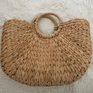 Beige flätad shoppingväska med runda handtag - Säljer en stor och rymlig strandväska i beige flätat material. Väskan har två runda handtag och en mjukt bågformad siluett. Perfekt för att bära med sig allt man behöver till stranden eller stan. Enkel och stilren design som passar till många outfits.