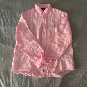 Rosa linneskjorta från Polo Ralph Lauren - Ljusrosa skjorta från Polo Ralph Lauren i luftig linnekvalitet. Klassisk button-down krage, långa ärmar och broderad blå logga på bröstet. Perfekt för en chill och stilren look, lätt att matcha med jeans eller shorts. Men behöver strykas så är den som ny. Passar runt längden 160.