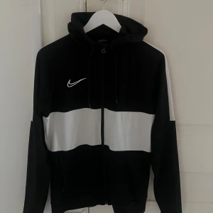 Svartvit Nike hoodie med dragkedja - Snygg svart och vit hoodie från Nike med dragkedja framtill och huva med snörning. Tröjan har en vit rand över bröstet och ärmarna, samt Nike-logga på bröstet. Perfekt för en sportig och avslappnad stil.