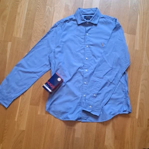 Blå skjorta från Polo Ralph Lauren - Snygg blå skjorta från Polo Ralph Lauren i slim fit-modell. Skjortan har klassisk krage, knäppning framtill och det ikoniska broderade polospelar-loggan på bröstet. Perfekt för dig som gillar stilrena och tidlösa plagg.