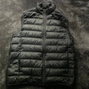 Svart dunväst från Uniqlo - Säljer en svart, quiltad dunväst från Uniqlo Ultra Light Down. Västen är lätt, har hög krage och dragkedja framtill. Perfekt för lager-på-lager och enkel att bära med sig. Klassisk och stilren design utan ärmar.