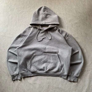 Grå hoodie från MODNI med huva - En stilren grå hoodie från MODNI med klassisk huva och snörning. Tröjan har en stor magficka och ribbade muddar vid ärmslut och nederkant. Perfekt för chill dagar och enkel att matcha med jeans eller joggers.