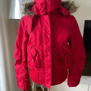 Röd bomberjacka med fuskpäls - Röd bomberjacka med huva och fuskpälsdetalj. Jackan har dragkedja och knappar framtill, stora fickor med både dragkedja och lock, samt ribbade muddar i midja och ärmslut. Perfekt för kyliga dagar och riktigt snygg färg som sticker ut.