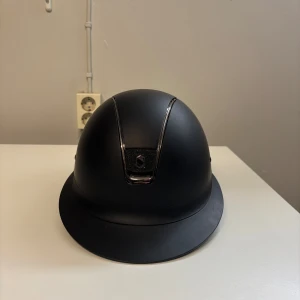 Svart ridhjälm från Samshield - Snygg svart ridhjälm från Samshield med matt finish och kromade detaljer framtill. Hjälmen har en klassisk rundad form och bred skärm, perfekt för dig som vill ha både stil och säkerhet i stallet.