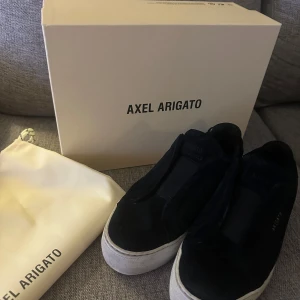 Svarta sneakers från Axel Arigato - Stilrena svarta sneakers från Axel Arigato i mocka med vit platt sula. Diskret guldtext på sidan och minimalistisk design. Skorna har rund tå och levereras med originalkartong och dustbag. Perfekta för dig som gillar en clean och modern look.
