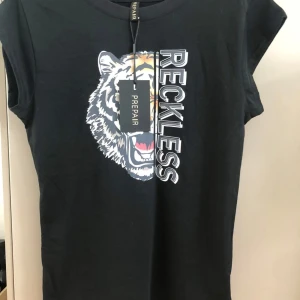 Svart t-shirt med tigertryck från Prepair - Svart t-shirt från Prepair med coolt tigertryck och texten 'RECKLESS' framtill. Modellen är kortärmad med rund hals och har en normal passform. Materialet är mjuk bomull som känns skönt mot huden. Perfekt för dig som gillar statement-plagg med attityd.