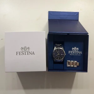 Festina klocka med blå urtavla - Stilren Festina herrklocka med blå urtavla och datumvisning. Klockan har ett klassiskt runt silverfärgat boett och länkarmband i metall. Levereras i originalask med extra länkar för perfekt passform.