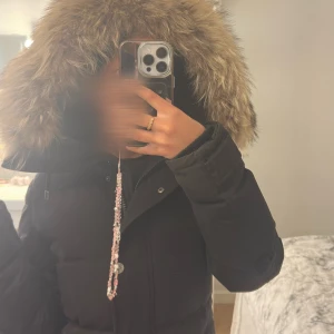 Hollies Subway jacka - Snygg svart dunjacka från Hollies med stor fluffig pälskantad huva. Jackan har långa ärmar, dragkedja och knappar framtill samt markerad midja. Perfekt för kalla dagar och ger en riktigt stilren look.