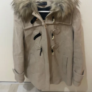 Beige parkajacka med fuskpäls - Snygg beige parkajacka från H&M med stor huva och fluffig fuskpäls. Jackan har dragkedja och dekorativa knäppen framtill samt två fickor. Perfekt för kyliga dagar och har en klassisk, tidlös look.