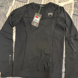 Svart tight tränings-tröja från Venum - Köpt från Zalando helt ny oanvänd för 609kr och säljer den nu för snabb affär. Svart långärmad tränings-tröja från Venum med tight passform och sportig design. Tillverkad i stretchigt syntetmaterial som andas, perfekt för gym eller kampsport. Snygg Venum-logga på bröstet och diskreta sömdetaljer för extra stilpoäng.