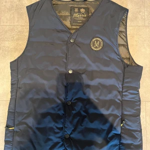 Morris marinblå dunväst - Snygg marinblå dunväst från Morris med klassisk passform. Västen har knäppning framtill, rund halsringning och ett broderat Morris-emblem på bröstet. Den är sparsamt använd och i mycket bra skick. Tidigare såld på CareofCarl: https://www.careofcarl.se/sv/morris-bridge-gilet-old-blue