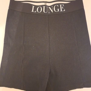 Svarta shorts från Lounge - Svarta shorts från Lounge med bred elastisk midja och vit logga framtill. De är ribbade och har en clean, enkel look som är både chill och snygg.                                                            Passformen är normal och materialet känns mjukt mot huden, perfekt för att hänga hemma eller dra på sig till träningen.