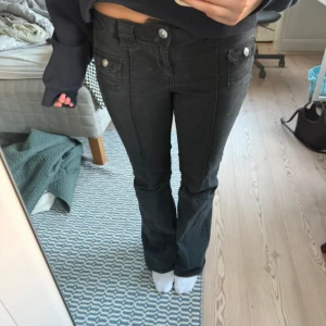 Svarta bootcut jeans - Snygga svarta jeans med bootcut passform och coola fickdetaljer framtill. Byxorna har knappar på fickorna och markerade sömmar längs benen som ger en schysst look. Perfekta för dig som gillar en lite retro vibe och vill ha jeans som sticker ut.