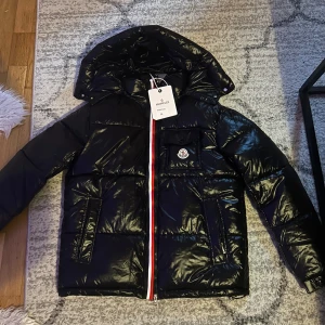 Svart Moncler pufferjacka med huva - Säljer min Moncler Maya-jacka Tyvärr passar inte storleken som jag hade önskat, så jag säljer den vidare. Jackan är helt ny och oanvänd. Den har QR-kod för äkthetskontroll. Perfekt för dig som vill ha en stilren och varm dunjacka av hög kvalitet!