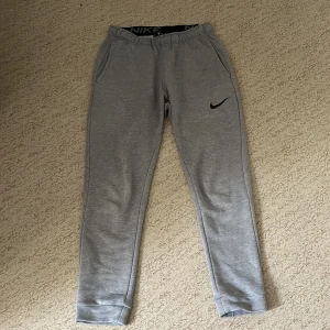 Grå Nike Dri-FIT mjukisbyxor M - Snygga grå mjukisbyxor från Nike med svart swoosh-logga på benet. Byxorna har elastisk midja, sidofickor och är tillverkade i Dri-FIT-material som andas. Perfekta för chill eller träning, standard fit och riktigt sköna att ha på sig.