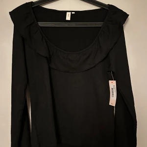 Svart blus med volangdetaljer - Svart blus  med bred volang runt ringningen och volang i ärmsluten. Långärmad modell i mjukt tyg med lätt struktur. Helt ny med prislapp