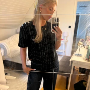 Svart t-shirt med silver knappar - Svart t-shirt med diskret rutigt mönster och silvriga dekorativa knappar på axeln. T-shirten har korta ärmar och en rak passform. Perfekt för dig som gillar stilrena detaljer och vill ha något som sticker ut lite extra.