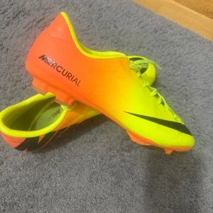 Nike Mercurial Victory IV FG fotbollsskor - Säljer ett par Nike Mercurial fotbollsskor i en riktigt fet färgkombination av neon-gult och orange med svarta detaljer. Skorna har snörning och är gjorda i syntetmaterial för bästa känsla på planen. Perfekta för dig som vill sticka ut på matchen!