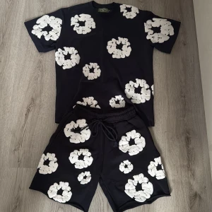 Svarta shorts med vita blommor Denim Tears - Svarta shorts från Denim Tears med stora vita blommor tryckta över hela plagget. Shortsen har elastisk midja med snörning och är tillverkade i ett mjukt material som känns skönt mot huden. Perfekta för en chill sommarlook.