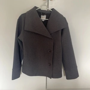 Grå kavajjacka från Gina Tricot - Snygg grå kavajjacka från Gina Tricot med bred krage och två knappar framtill. Jackan har en rak och lite boxig passform, långa ärmar och är gjord i ett mjukt ull-liknande material. Perfekt för dig som gillar stilrena och klassiska plagg.