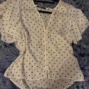 Vit prickig blus från Gina Tricot - Supersöt vit blus från Gina Tricot med svarta prickar, v-ringning och knappar hela vägen framtill. Blusen har korta, luftiga ärmar och lätt volang vid axlarna. Perfekt för dig som gillar en lekfull och trendig stil.