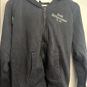 Svart hoodie Peak Performance - Svart hoodie från Peak Performance med dragkedja framtill och broderad logga på bröstet. Tröjan har huva med snörning och två stora fickor framtill. Perfekt för en avslappnad och sportig stil.