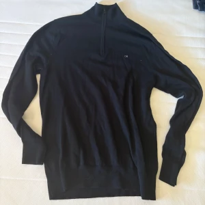 Svart half zip tröja från Calvin Klein - Svart långärmad tröja från Calvin Klein med half zip och liten CK-logga på bröstet. Tröjan har hög krage och är i ett mjukt, stickat material som passar perfekt till en clean och stilren look.