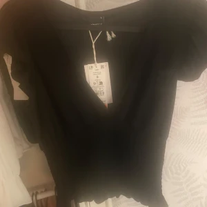 Svart v-ringad blus från Gina Tricot - Svart blus från Gina Tricot med v-ringning och korta ärmar. Toppen har smockad midja och lätt volangkant nertill, vilket ger en feminin och trendig look. Perfekt att matcha med jeans eller kjol för en snygg outfit.