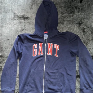 Marinblå hoodie med dragkedja från GANT - Snygg marinblå hoodie från GANT med stor röd logga framtill. Tröjan har huva, dragkedja och två fickor framtill. Tillverkad i mjuk bomull, perfekt för chill dagar. Klassisk och sportig look som alltid funkar.