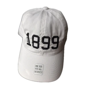 Vit keps från CCM med brodyr - Vit keps från CCM med svart broderad text 'EST 1899' framtill. Justerbar metallspänne baktill för perfekt passform. Klassisk böjd skärm och stilren look, tillverkad i bomull. One size fits all.
