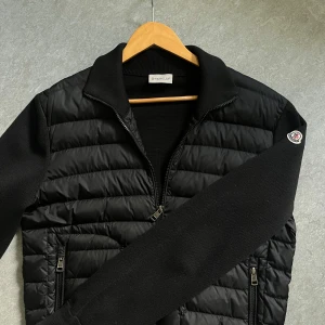 Moncler cardigan - Snygg svart cardigan från Moncler med quiltad framsida och ärmar i stickad bomull. Jackan har två fickor med dragkedja och klassisk Moncler-logga på ärmen. Perfekt för dig som vill ha en stilren och exklusiv look. Storlek M men passar även S, för mer frågor kom privat!