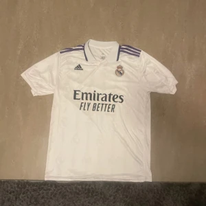Real Madrid Benzema tröja Adidas L - Säljer en officiell Real Madrid matchtröja från Adidas i storlek L med Benzema och nummer 9 på ryggen. Tröjan är vit med lila detaljer på axlarna, har korta ärmar och klassisk krage. Materialet är lätt och andas med ClimaCool-teknologi. Perfekt för dig som älskar fotboll och Real Madrid.