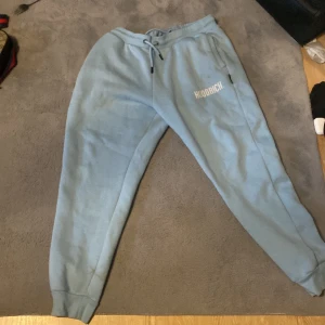 Ljusblå mjukisbyxor från Hoodrich - Säljer ett par ljusblå mjukisbyxor från Hoodrich med snörning i midjan och muddar vid bensluten. Byxorna har tryckt logga på ena benet och är tillverkade i mjukt material som är perfekt för chill eller streetwear.
