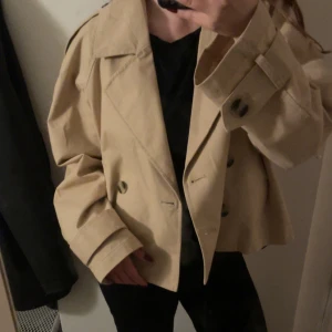 Beige trenchcoat från H&M Divided - Snygg beige trenchcoat från H&M Divided med dubbelknäppning, bred krage och axelklaffar. Jackan har raka fickor och en klassisk, lite boxig passform. Säljer den pga att den aldrig kommer till användning!
