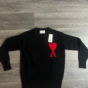 Svart stickad tröja från Ami Paris - Svart stickad tröja från Ami Paris med röd hjärtformad A-logga på bröstet. Klassisk rund halsringning, ribbade muddar och nederkant. Perfekt för dig som gillar stilrena plagg med en twist. Materialet är mjukt och håller värmen.