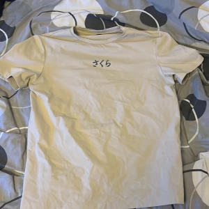 Beige t-shirt med japanskt tryck - Snygg beige t-shirt med japanska tecken på bröstet och ett stort konstnärligt tryck på ryggen som visar en samuraj under ett blommande träd med röd sol i bakgrunden. T-shirten har rund hals och korta ärmar, perfekt för dig som gillar unik streetstyle med asiatiska vibbar.