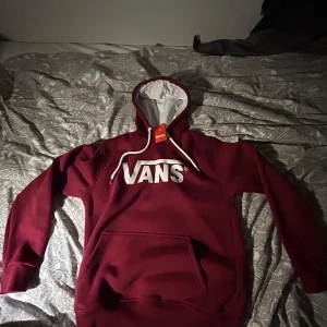 Vinröd hoodie från Vans med logga - Snygg vinröd hoodie från Vans med stor vit logga på bröstet. Tröjan har huva med vita snören. Insidan av huvan är vit och materialet känns mjukt och skönt, perfekt för chill dagar.