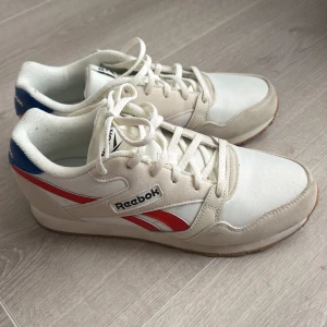 Vita Reebok sneakers med röda detaljer - Snygga vita Reebok-sneakers med röda och blå detaljer. Klassisk retromodell med rund tå, snörning och platt sula. Använda endast en gång och insåg att jag köpte fel storlek.