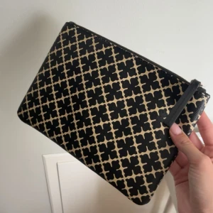 By malina clutch - Clutch väska i svart och beige med ett grafiskt rutigt mönster. Väskan har dragkedja upptill och är tillverkad i ett slätt syntetmaterial. Perfekt att bära i handen eller lägga i en större väska för att organisera småsaker.